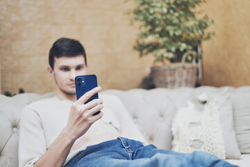 guy sitting on couch holds smartphone in his hand, online shopping and chatting with friends concept