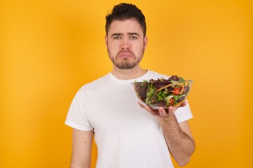 young handsome Caucasian man holding a salad bowl against yellow wall crying desperate and...