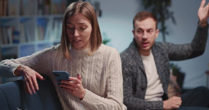 Angry caucasian sad young man and woman making up quarrel in their apartment. Emotional negative couple agruing and using mobile phones separately.