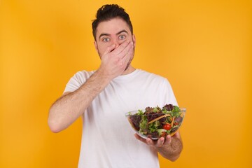 Upset young handsome Caucasian man holding a salad bowl against yellow wall, covering her mouth with both palms to prevent screaming sound, after seeing or hearing something bad.