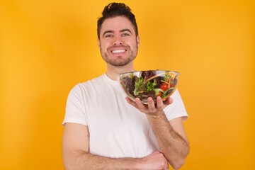 Cheerful young handsome Caucasian man holding a salad bowl against yellow wall with hand near face. Looking with glad expression at the camera after listening to good news. Confidence.