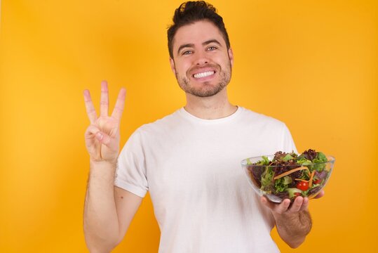 Young Handsome Caucasian Man Holding A Salad Bowl Against Yellow Wall Showing And Pointing Up With Fingers Number Three While Smiling Confident And Happy.