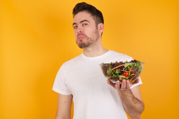 young handsome Caucasian man holding a salad bowl against yellow wall with snobbish expression...