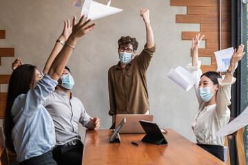 Smiling business people group,teamwork of young asian,caucasian throwing papers, documents, wearing face mask having fun, happy in office, workplace after project is done. Celebrating success concept.