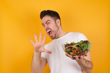 Dissatisfied young handsome Caucasian man holding a salad bowl against yellow wall frowns face, has disgusting expression, shows tongue, expresses non compliance, irritated with somebody.