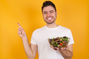 Positive young handsome Caucasian man holding a salad bowl against yellow wall with beaming smile pointing with two fingers and looking on empty copy space. Advertisement concept.