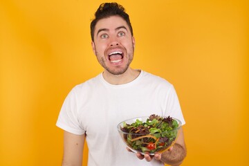 Surprised young handsome Caucasian man holding a salad bowl against yellow wall, shrugs shoulders, looking sideways, being happy and excited. Sudden reactions concept.