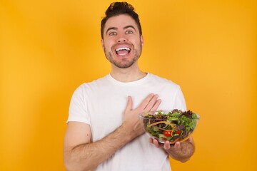 Happy smiling young handsome Caucasian man holding a salad bowl against yellow wall has hands on chest near heart. Human emotions, real feelings and facial expression concept.