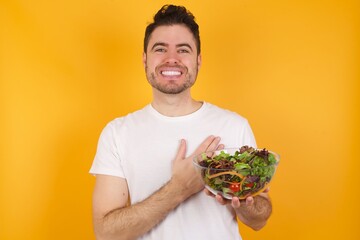 Honest young handsome Caucasian man holding a salad bowl against yellow wall keeps hands on chest,...