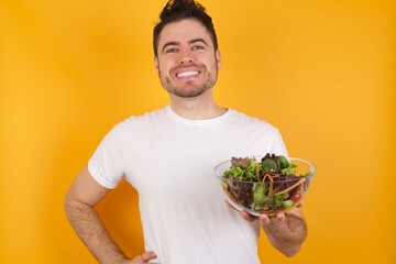 Studio shot of cheerful young handsome Caucasian man holding a salad bowl against yellow wall keeps hand on hip, smiles broadly.