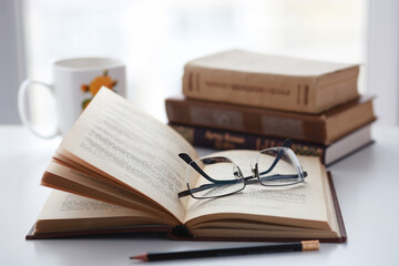 Old books, glasses, pencil and cup of coffee on white table background against window light with copy space