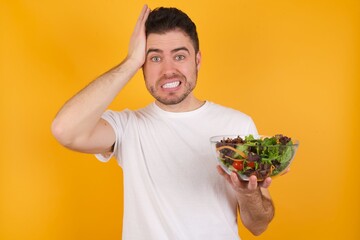 Portrait of confused young handsome Caucasian man holding a salad bowl against yellow wall holding...