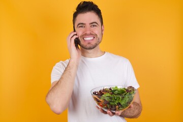 Portrait of a smiling young handsome Caucasian man holding a salad bowl against yellow wall talking on mobile phone. Business, confidence and communication concept.