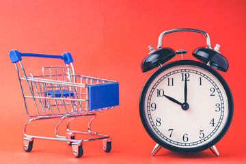 An empty shopping cart and an alarm clock on a red scarlet background. Shopping, online shopping concept, time to buy concept, copy space, top view,