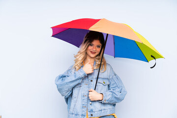 Teenager russian girl holding an umbrella isolated on blue background with thumbs up because something good has happened