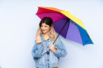 Teenager russian girl holding an umbrella isolated on blue background inviting to come with hand. Happy that you came