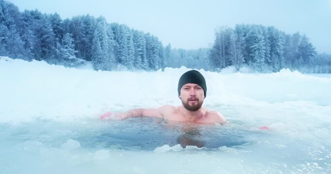 Winter swim, cold treatment. Young man swims in the icy water with snow covered forest on the background