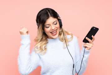 Teenager girl isolated on pink background listening music with a mobile making rock gesture
