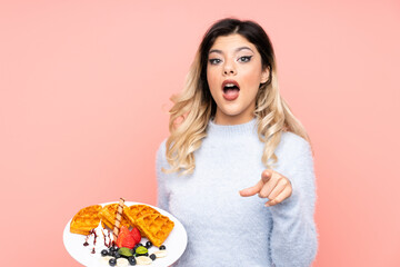 Teenager girl holding waffles on isolated pink background surprised and pointing front