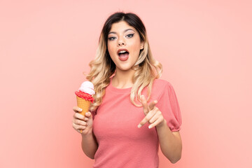 Teenager girl holding a cornet ice cream isolated on pink background surprised and pointing front