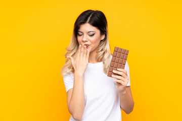 Teenager girl isolated on  yellow background taking a chocolate tablet and surprised