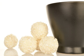 Several round chocolates with coconut flakes with a ceramic black cup, close-up, isolated on white.
