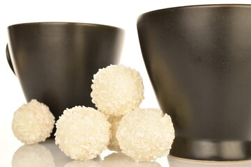 Several round chocolates with coconut flakes with two ceramic black cups, close-up, isolated on white.