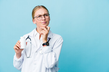 Young russian doctor woman holding pills bottle on blue looking sideways with doubtful and skeptical expression.