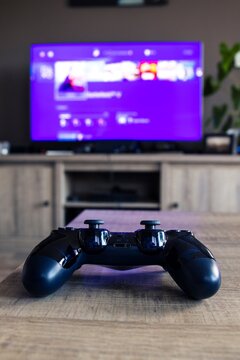 A vertical portrait of a sony playstation 4 controller in front of a television with the playstation home menu displayed on it. The PS4 video game controller is turned on and ready to be played with.
