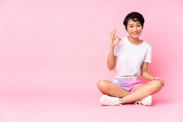 Young Vietnamese woman with short hair sitting on the floor over isolated pink background showing ok sign with fingers