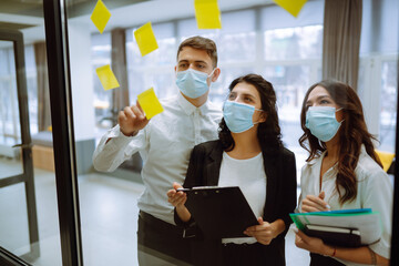 Three business colleagues in protective face mask writing on stickers on office glass board. Office workers are discussing and planning during pandemic coronavirus. Startup, planning, management.