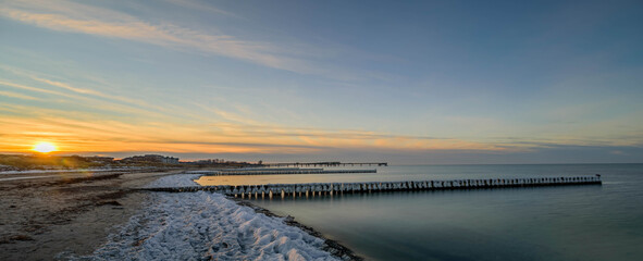 Winter landscape on the Baltic sea with the beach, rows of frozen wooden posts, sea bridge and hotels. Sunset by Heiligenhafen in winter with snow and frost, Schleswig-Holstein, Germany. 