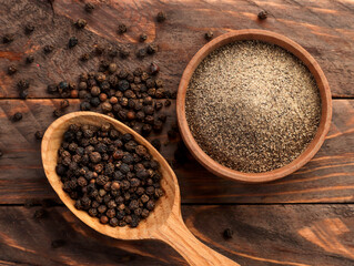Black peppercorns in a spoon and black ground pepper in a plate on a wooden background. Top view