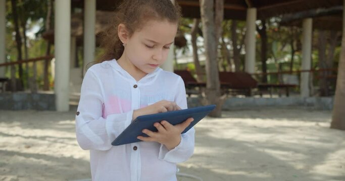 Curious cute girl 7-8 years old is having fun with a digital tablet device watching a cartoon while walking on the beach. The child has a rest plays in the tablet outdoors.