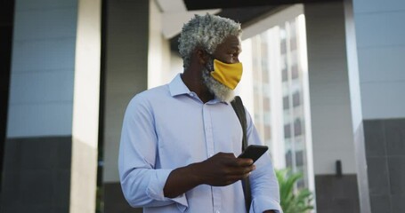African american senior man wearing face mask using smartphone and looking around in corporate park - Powered by Adobe