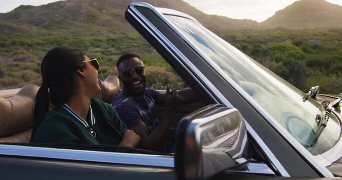 Diverse couple driving on sunny day in convertible car raising their arms in the air and smiling