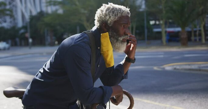 African american senior man wearing face mask talking on smartphone while leaning on his bicycle on 