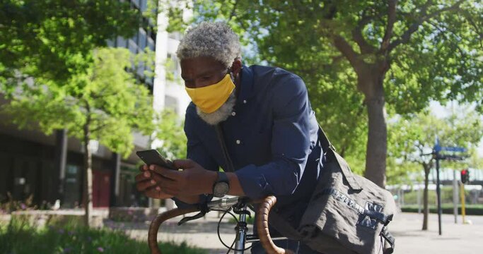 African american senior man wearing face mask using smartphone while leaning on his bicycle on the r