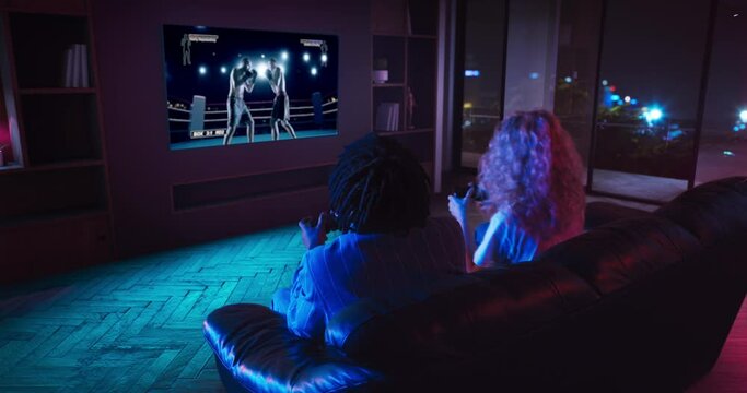 Young man and woman are playing fighting video game on TV. They are sitting on the sofa in the living room with red and blue neon lighting. Girl wins and celebrates victory.