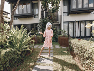 Picture of a lovely woman looking at side in white bathrobe and with towel on her head standing and posing on the garden at the background of villa. Summer concept
