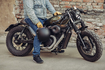 Obraz na płótnie Canvas Attractive biker in denim clothes holding a black helmet in his hand while sitting on a motorcycle with tape cross over optic. Brutal bike rider. Courage concept