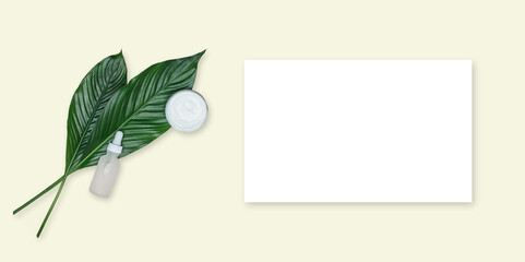 White card mockup with green leaf of peace lily - spathiphyllum, face cream and serum bottle on yellow background. Cosmetic background. Overhead view