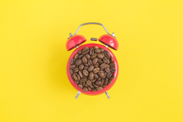 Top view of coffee beans on the dial of the red alarm clock in the center of the yellow background