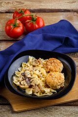 Pasta in a creamy sauce with mushrooms and chicken meatballs in a plate on a board on a blue napkin next to a tomato.