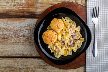 Pasta in a creamy sauce with mushrooms and chicken meatballs in a black plate on a wooden table on a napkin near the fork.
