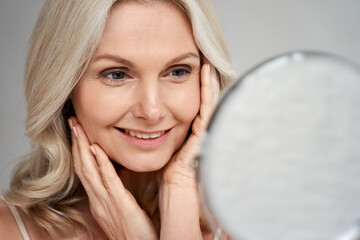 Happy 50s middle aged woman model touching face skin looking in mirror. Smiling mature older lady...