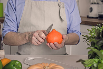 Man without face preparing pumpkin dish, cutting raw orange pumpkin with knife on kitchen table,...