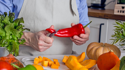 Man preparing a vegan dish of pepper, holding a pod of red pepper in his hands and cutting it with a knife on the kitchen table, the concept of home cooking healthy food from raw vegetables
