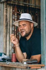 Portrait of an attractive adult and focused man with a beard and a black and white panama hat on his head, holding a cigar in his teeth, leaning his logs on the table. Relaxion concept.