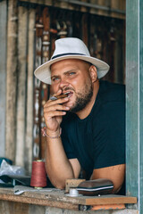 Portrait of a serious concentrated brutal man with a beard and a black and white panama hat on his head, holding a cigar in his teeth, leaning his logs on the table. Vacation concept.